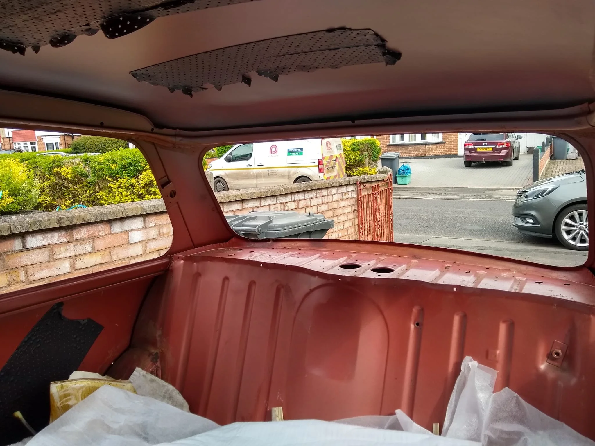 Looking out from inside an older, red car's trunk space, with the rear hatch open, towards a driveway with parked vehicles, bushes, and houses in the background.