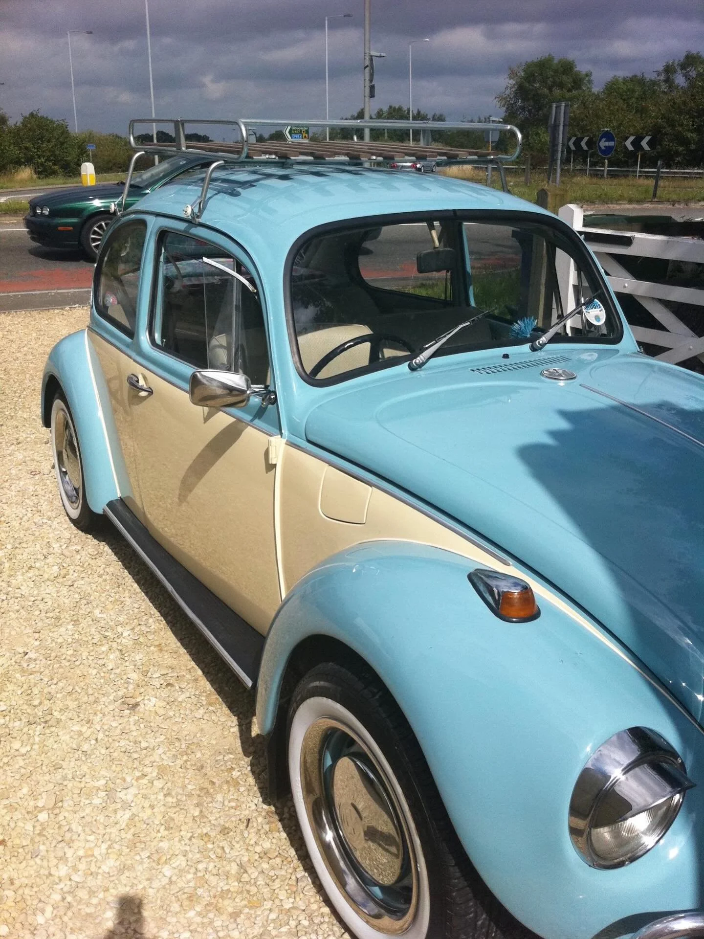 A light blue vintage Volkswagen Beetle parked on a gravel surface near a white wooden fence, with a green car and a cloudy sky in the background.