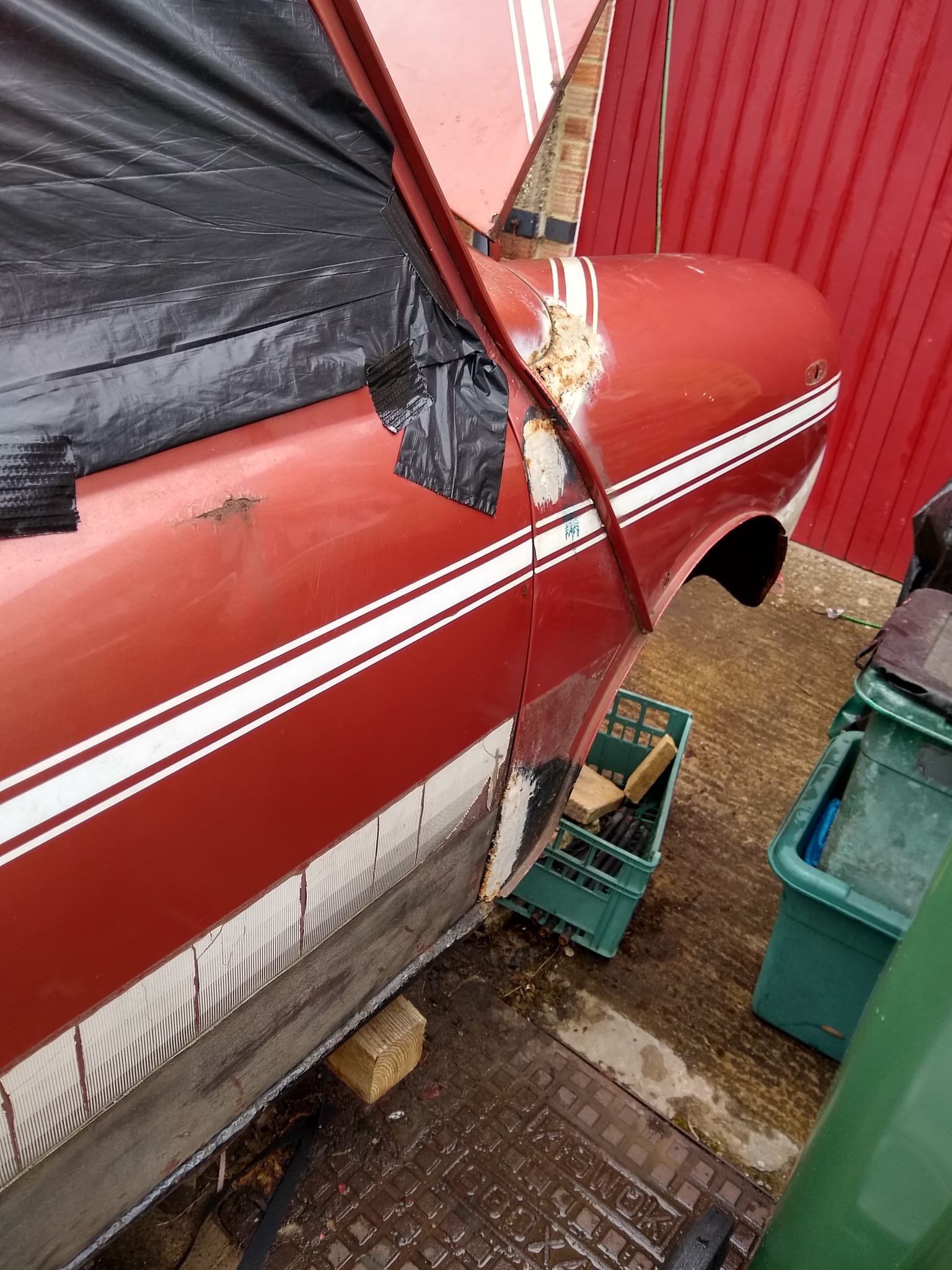 Close-up of a red vintage car with visible rust and damage, inside a workshop with tools and bins around.