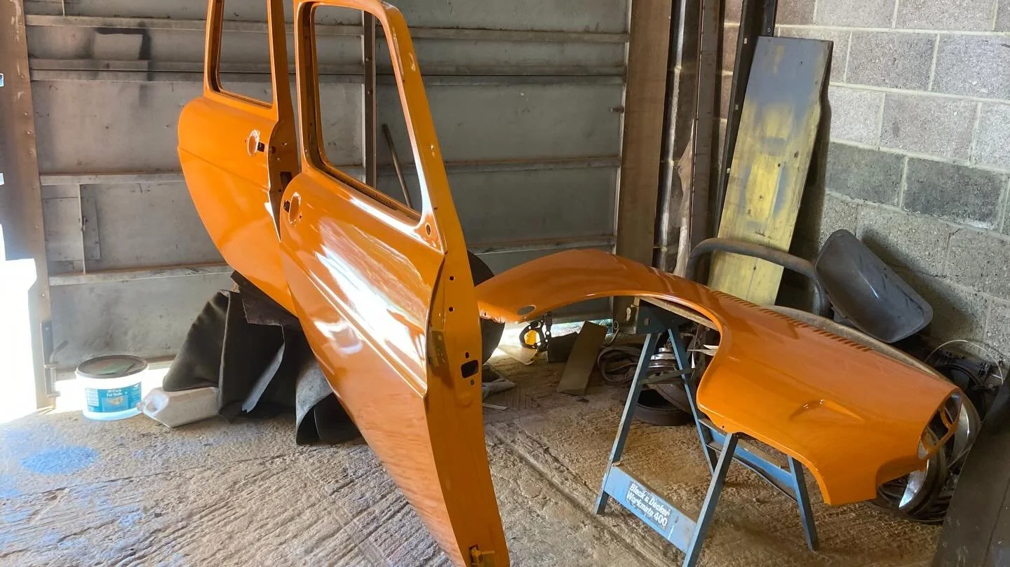Orange vintage car body parts in a garage, including doors and fenders, with some tools and supplies around.