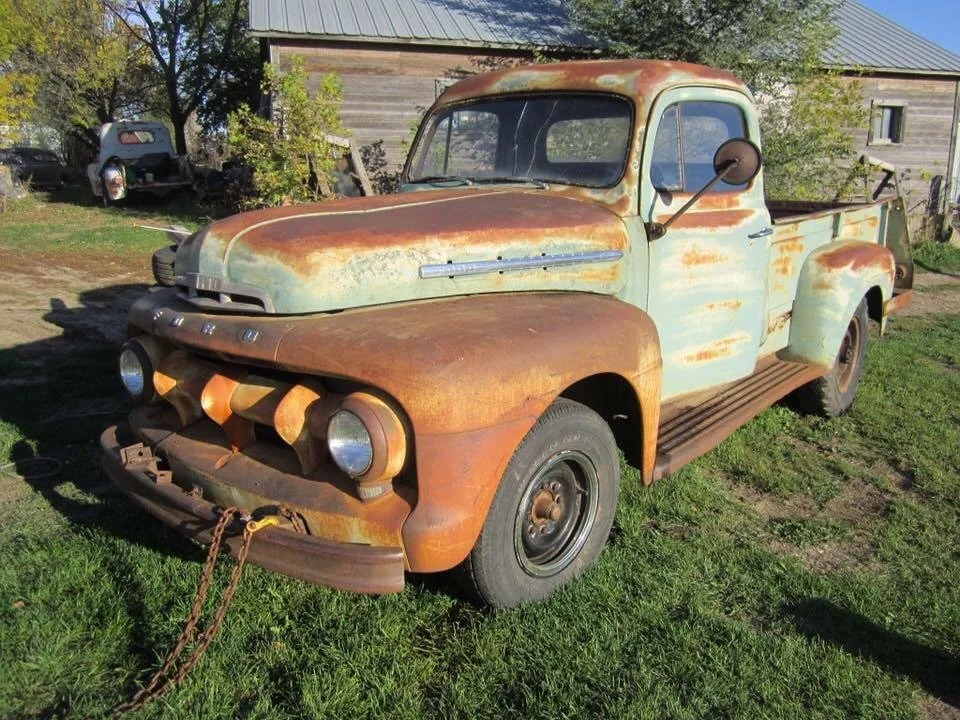 Old, rusty pickup truck parked on grass in front of a barn and trees.