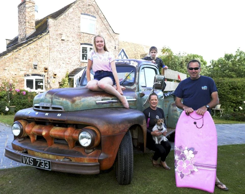 Four people, a woman, a man, and two children, with a small dog, standing near a rusty, vintage Ford truck, in front of a brick house during daytime.