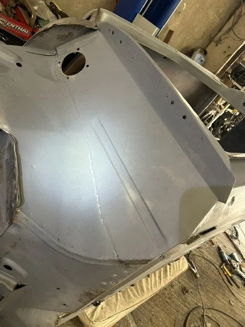 Metal boat hull in a workshop, showing a smooth interior surface with some weld marks and a round hole near the top left corner.