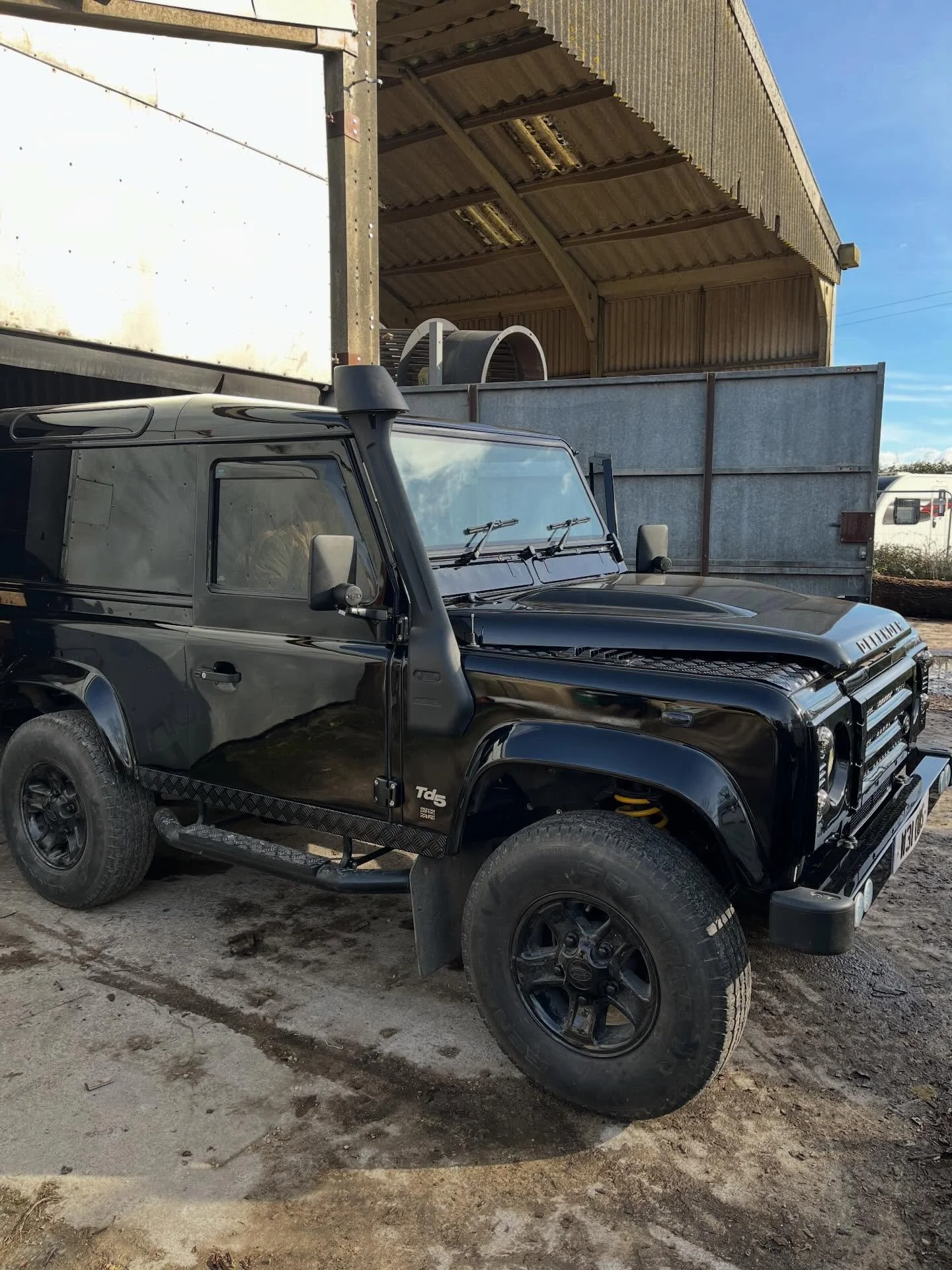 A black Land Rover Defender parked outside a warehouse with metal siding and a curved roof, under a clear blue sky.