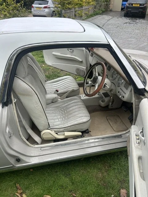 Interior of a vintage silver car with white seats, a brown steering wheel, and a dashboard with various controls, parked on a grassy area with other cars in the background.