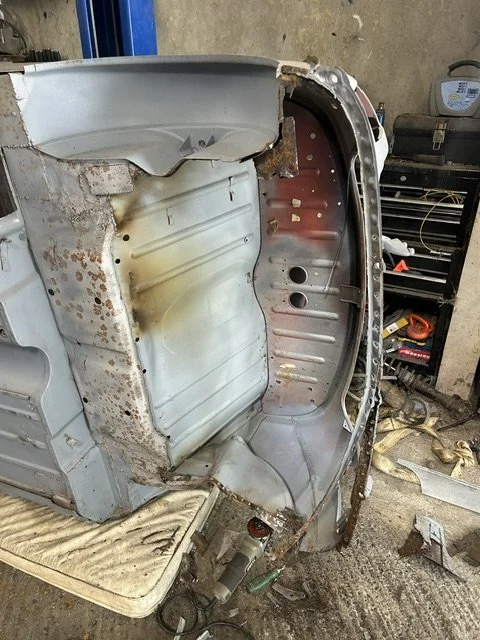 Rusted and partially disassembled metal vehicle body, possibly a car, in a workshop with tools and equipment visible in the background.