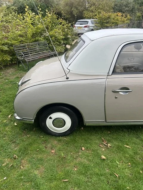 A small vintage silver car parked on grass with a shrub, empty garden bench, and other vehicles in the background.