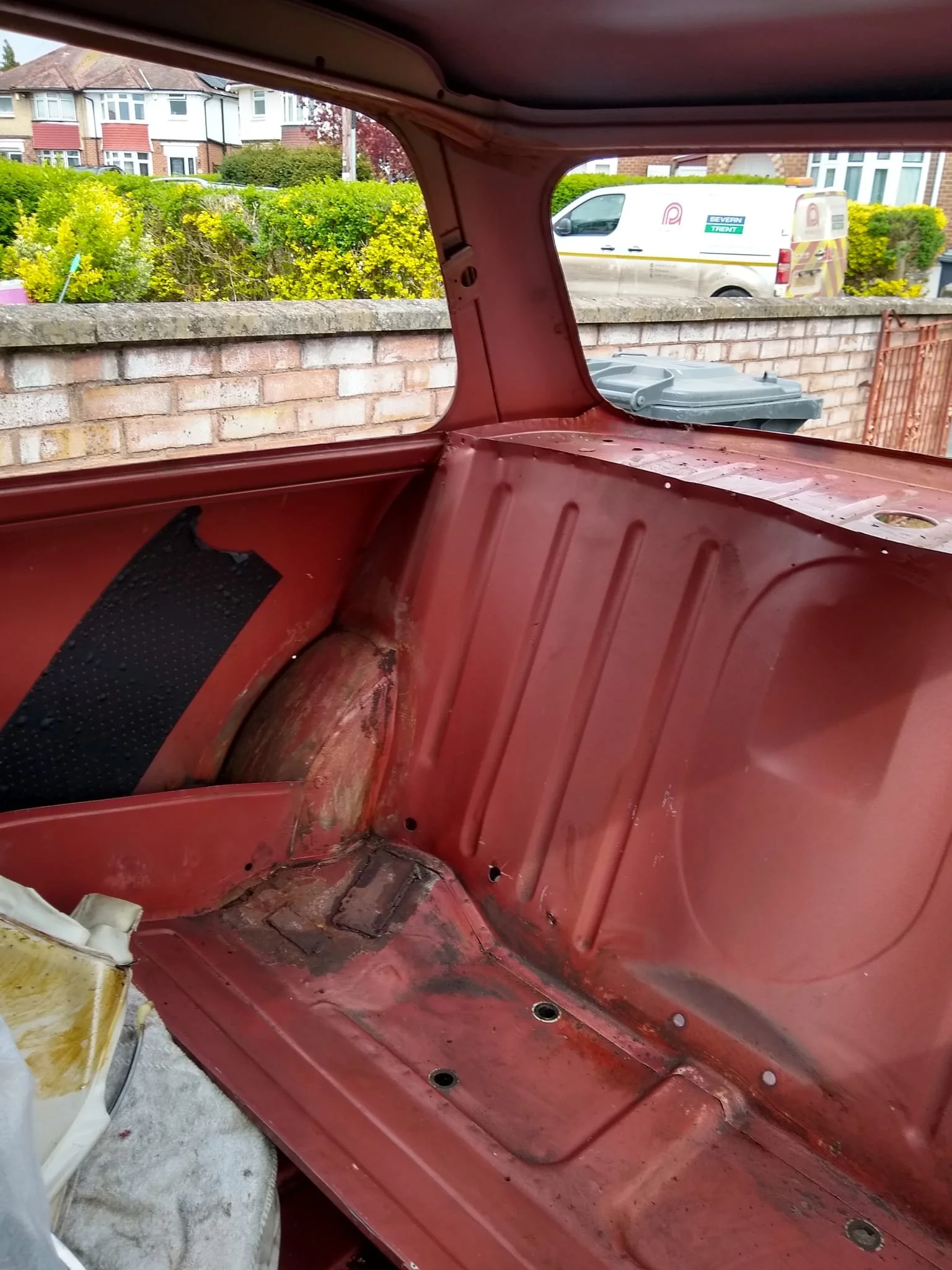 Interior of an old, empty red vehicle with visible rust and damage, looking out through the rear window onto a residential street with houses, a hedge, a parked white van, and some tools or equipment inside.