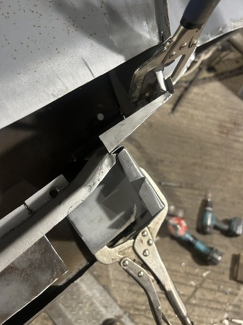 Close-up of a metalworking or assembly process with clamps securing metal pieces, with power drills and tools on a wooden work surface in the background.