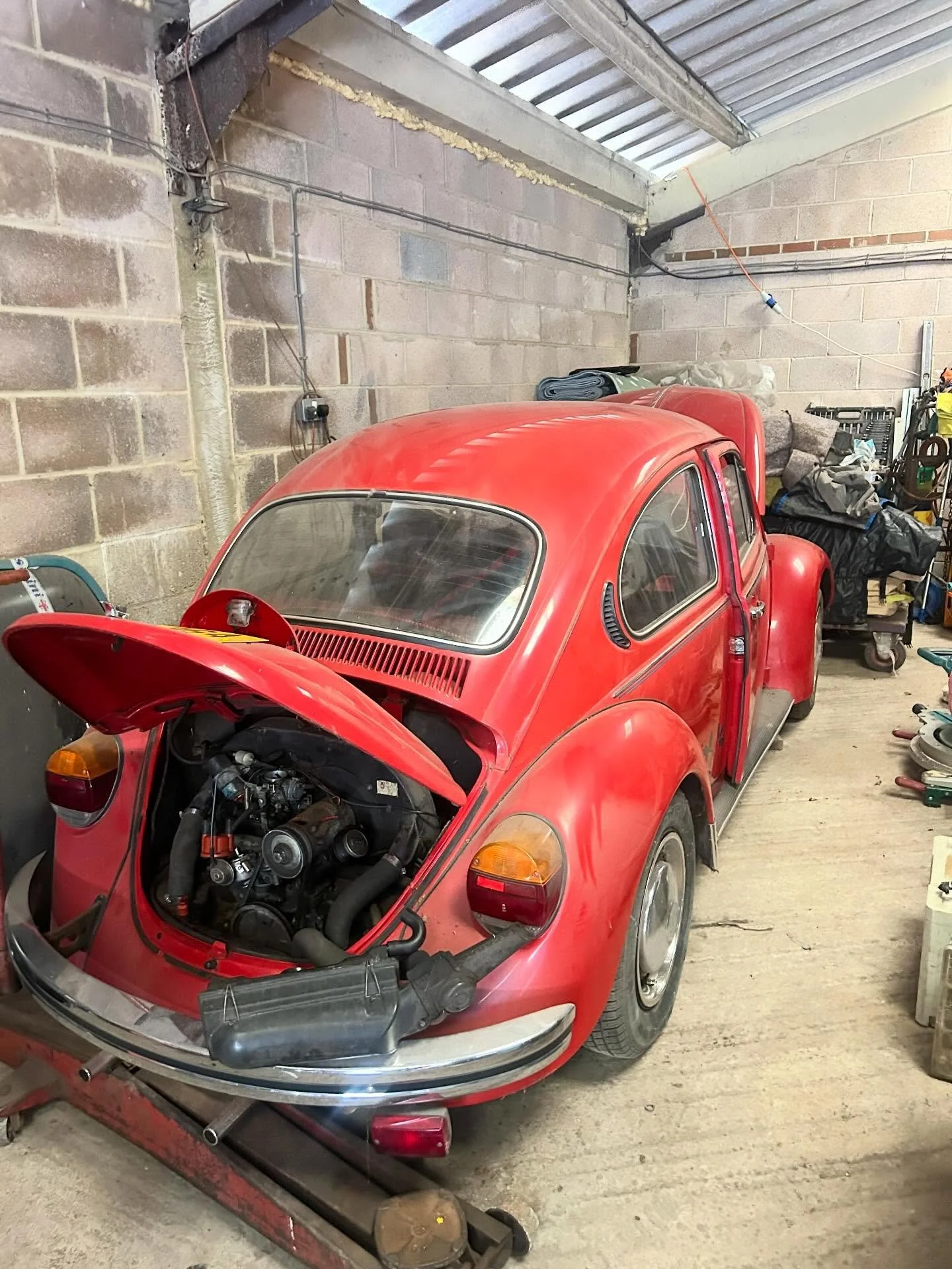A red vintage Volkswagen Beetle car in a garage with its engine hood open. The garage has unfinished brick walls, various tools, and supplies on shelves.