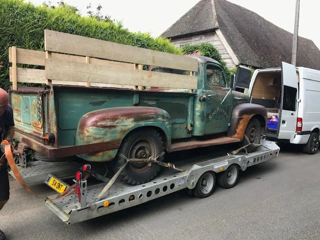 A vintage green pickup truck with a wooden flatbed loaded onto a trailer, parked on a residential street near a white van and a house with a shingled roof.