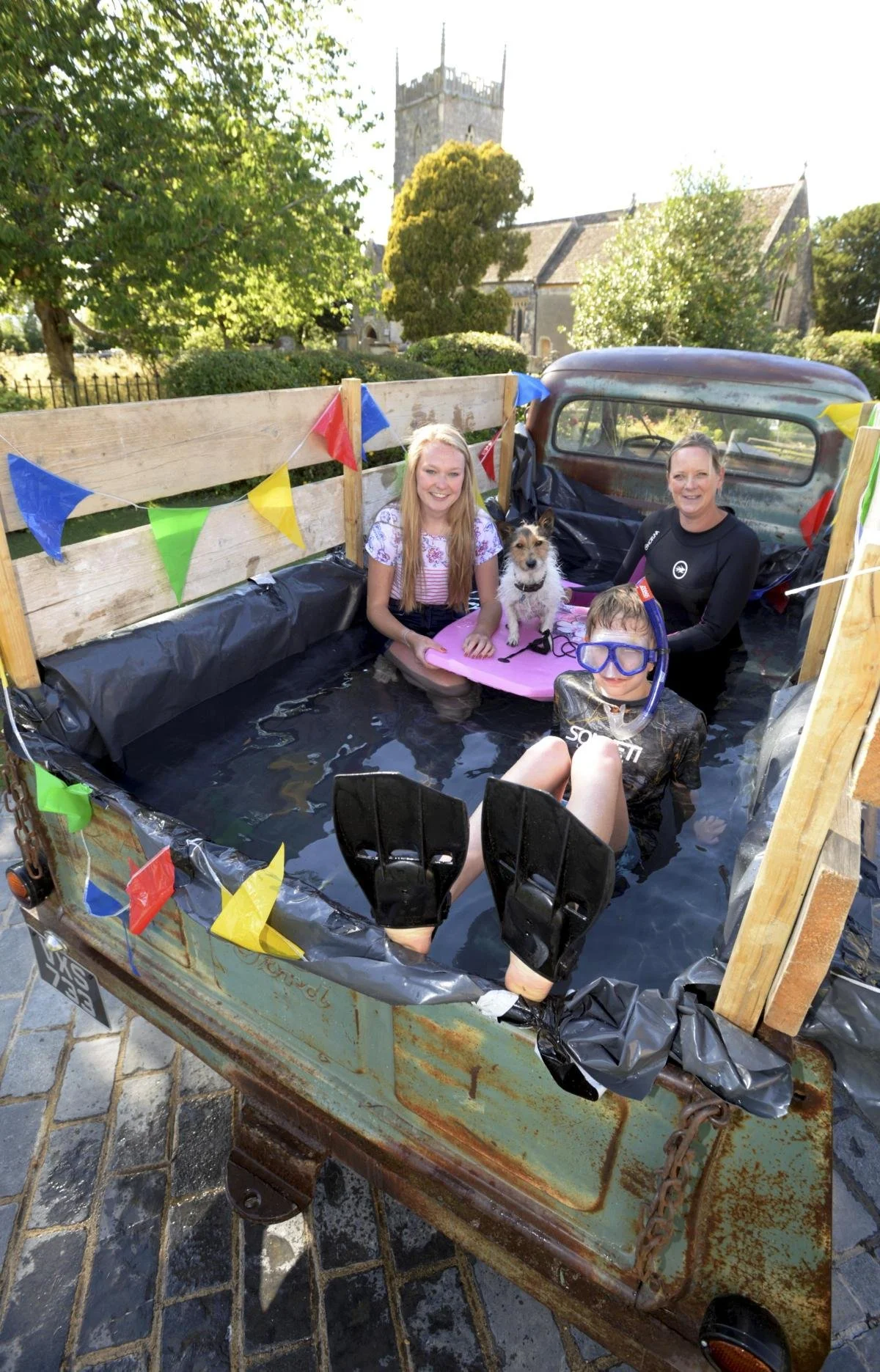 Three people and a dog sitting in a small, makeshift pool in the back of an old truck. The truck is decorated with colorful pennant banners. They are outdoors near trees and a church.