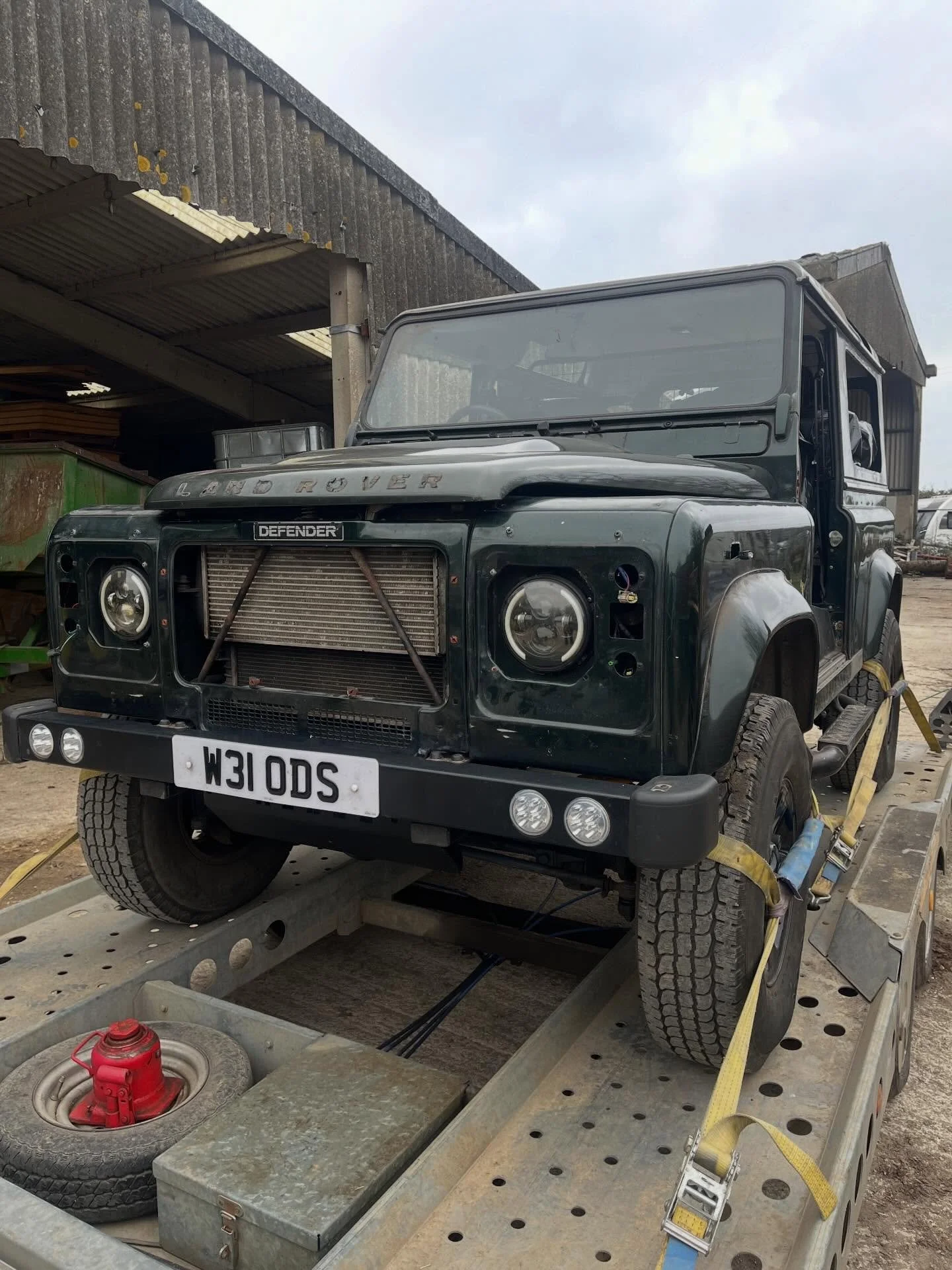 A black Land Rover Defender vehicle with a missing front grille, positioned on a flatbed trailer, with a grey building structure in the background and a partly cloudy sky.