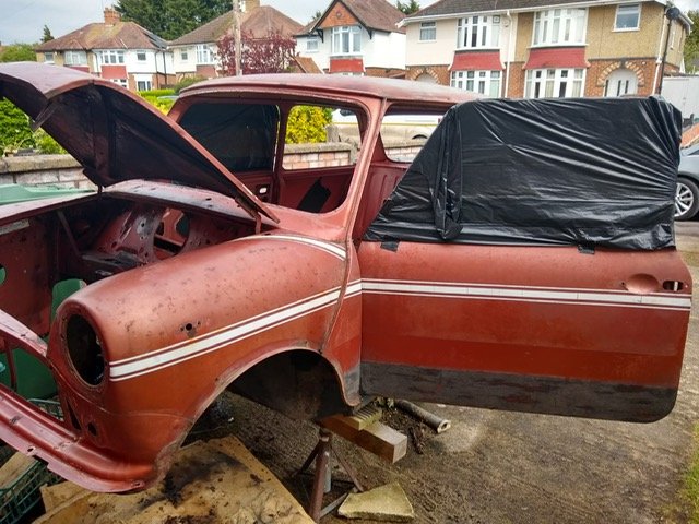 An old, rusty pickup truck with its hood open and one door covered with a black plastic sheet, missing its front wheels and resting on blocks in a residential area.