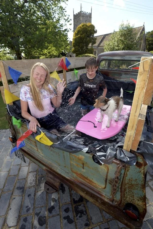 Two children and a puppy in a makeshift swimming pool set up in the back of an old rusty truck, with colorful bunting flags, on a brick-paved patio, with a church tower and trees in the background.