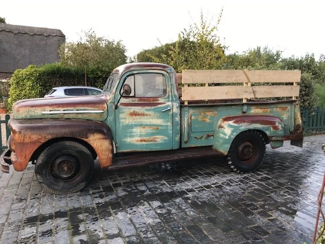 Old, rusted pickup truck with a wood bed parked on a cobblestone driveway, with greenery in the background.