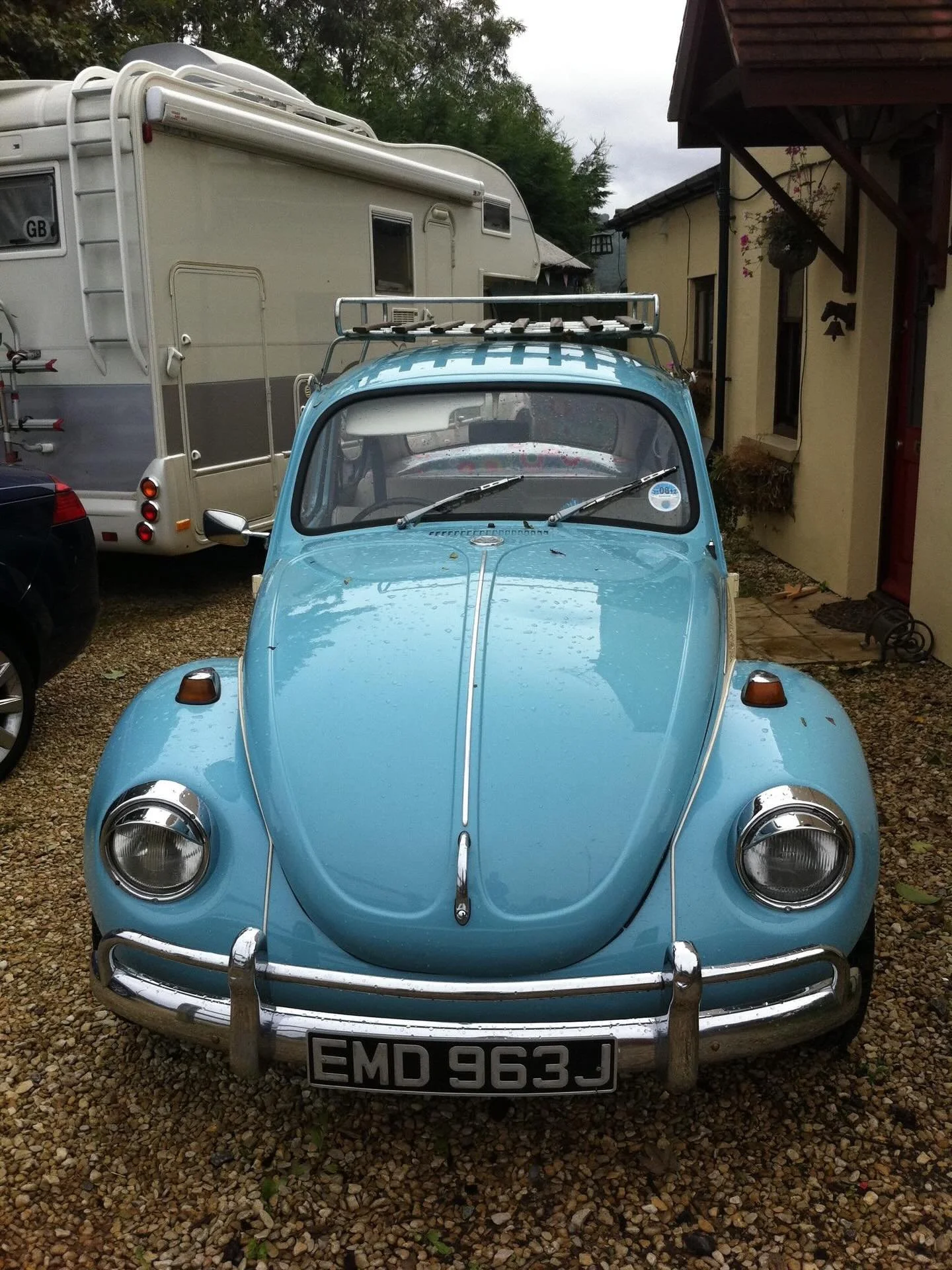 Front view of a vintage light blue car with a roof rack, parked on a gravel driveway in front of a house and neighboring camper trailer.