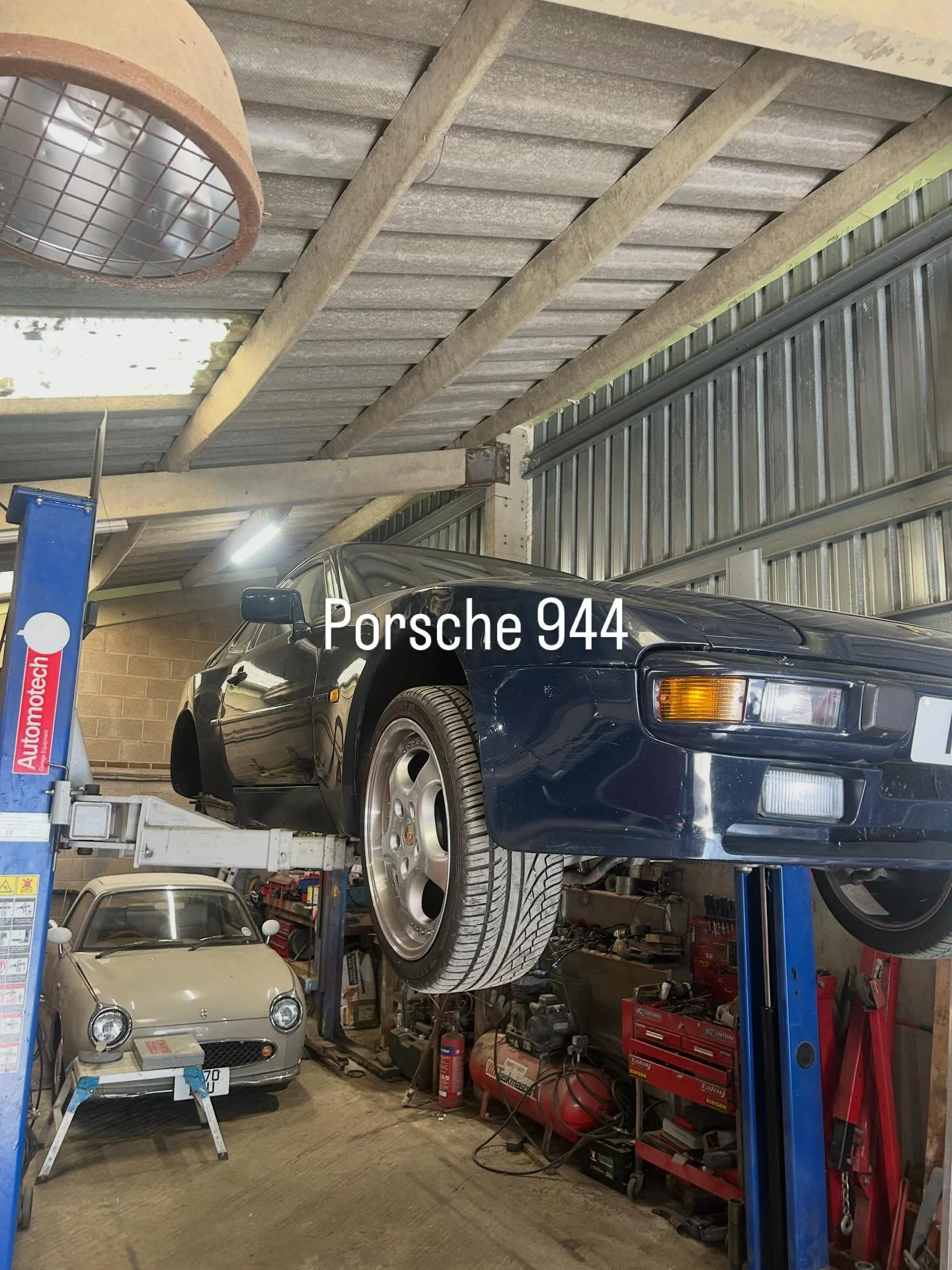 A garage with two Porsche cars, one black and one beige, inside a car lift. The garage has tools and equipment, including a red tool chest, and a fluorescent light. The black Porsche 944 is elevated on the lift, and the beige Porsche is parked on the