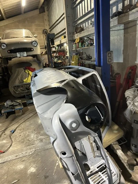 A car bumper resting on the floor in a garage or repair shop with tools and other car parts around, and another vehicle elevated on a lift in the background.