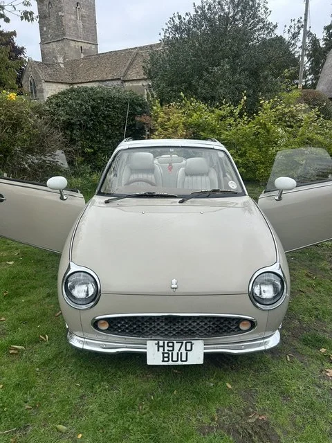 Front view of a vintage silver car with open doors, parked on grass, with a church tower and trees in the background.