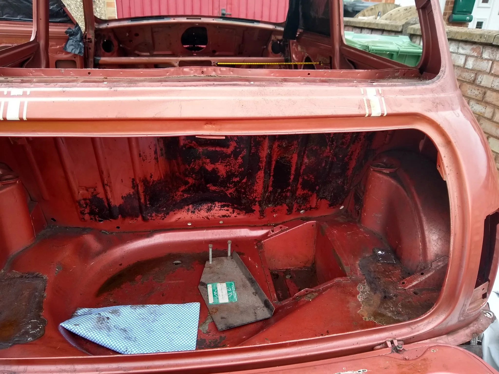 An old red vehicle stripped of its interior, showing the empty dashboard and cabin floor with blackened areas, a blue checkered cloth, and a green plastic container outside in the background.