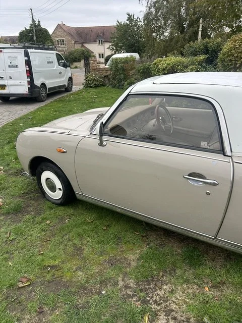 A vintage cream-colored car parked on a grassy area near a street, with modern vehicles and houses in the background.
