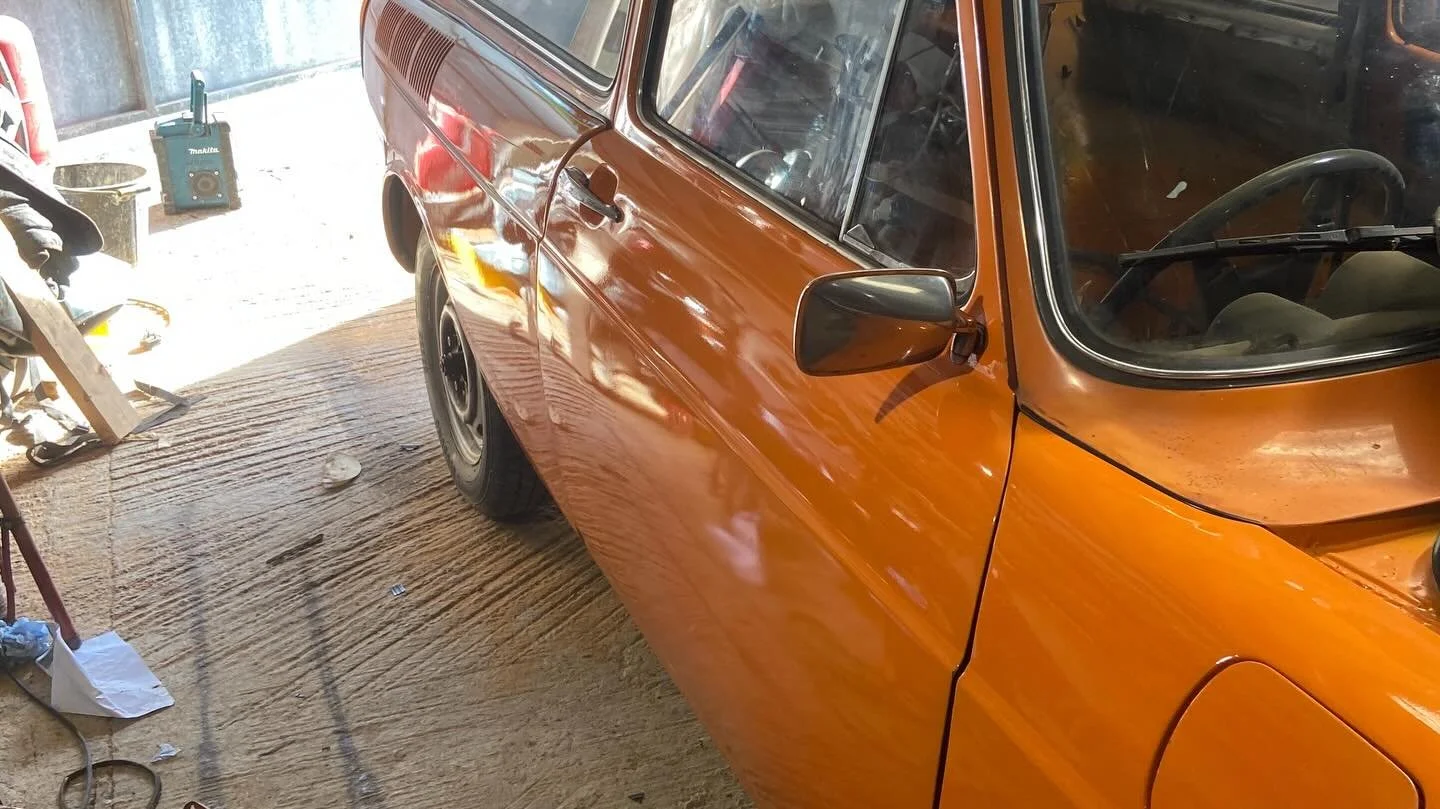 Rear side view of an orange vintage car inside a garage, with a damaged left front fender and a black tire.