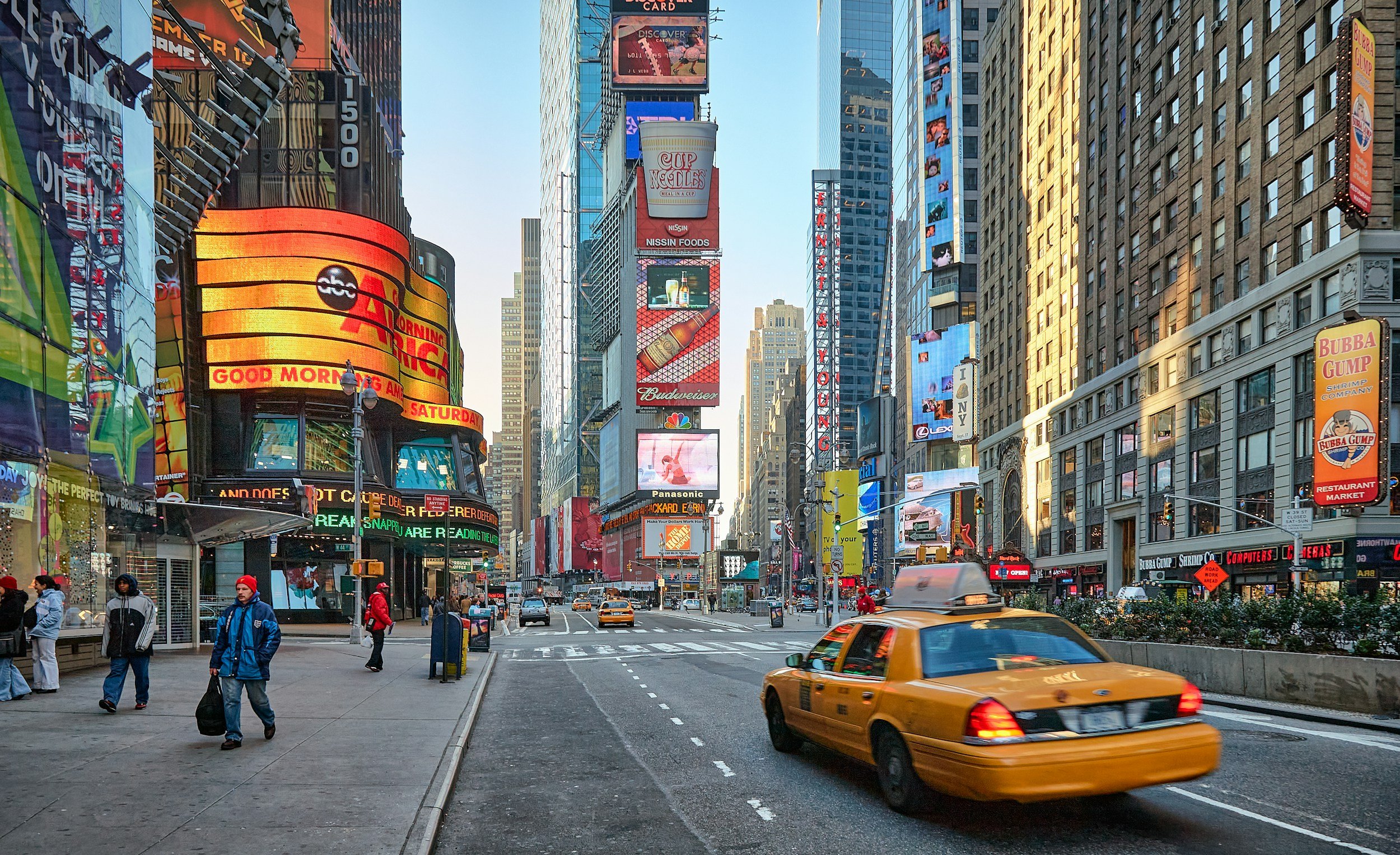 Times Square in New York City with tall buildings, bright digital billboards, and pedestrians. A yellow taxi is driving on the street.