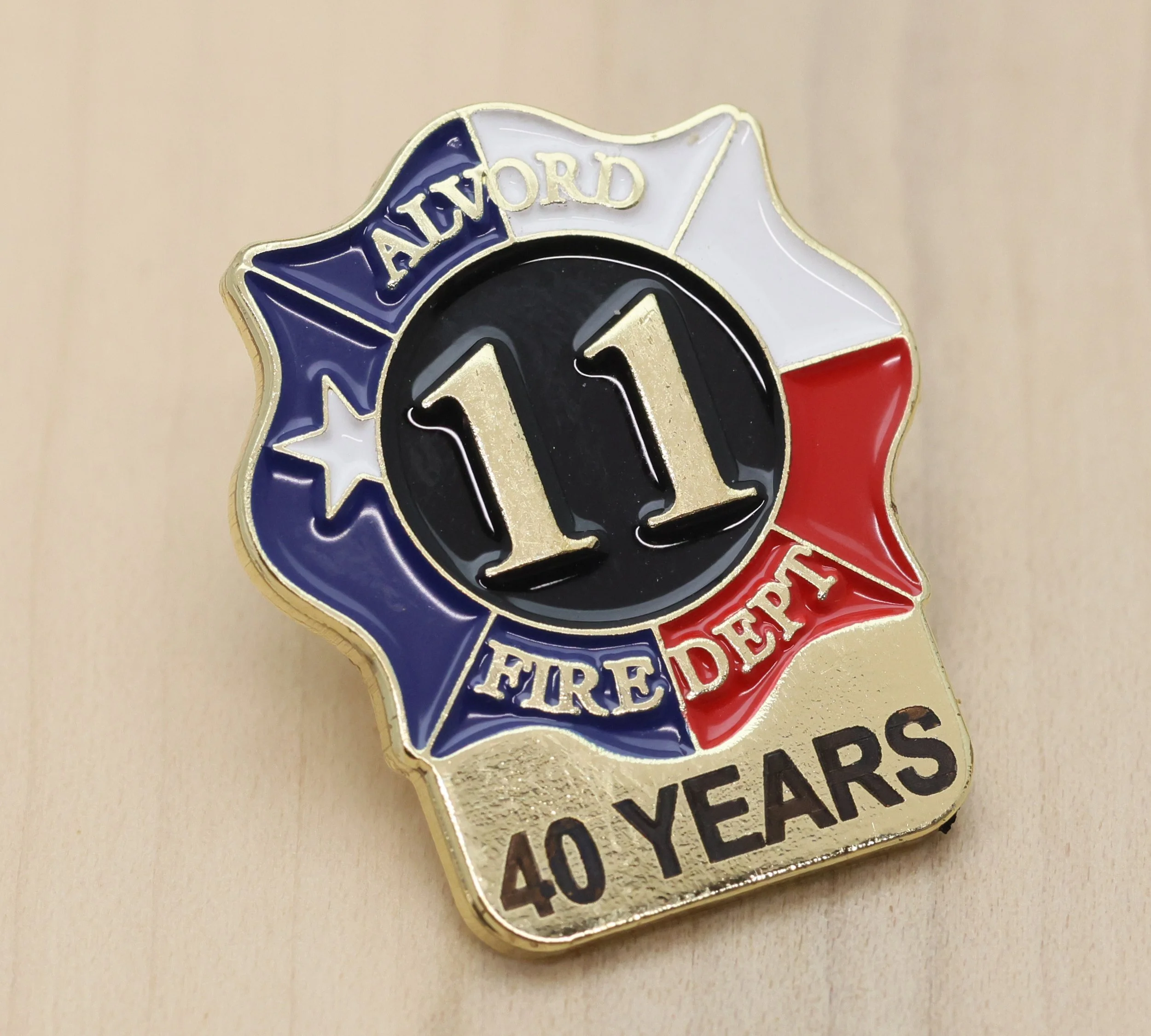Close-up of a years of service pin featuring a helmeted firefighter's helmet, with the words 'ALCOV' and 'FIRE DEPT' on a ribbon banner above and below the helmet, respectively, with red, white, and blue colors.