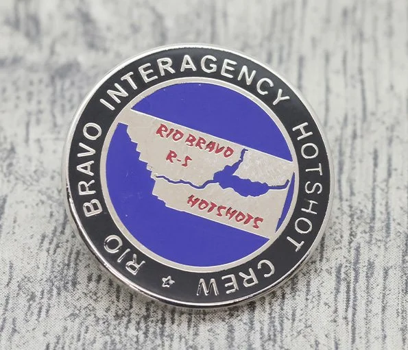 A circular service pin with a black border and white text reading 'BRAVO INTERAGENCY HOT SHOT CREW'. Inside, a regional map with red text 'RIO BRAVO R-S' and 'SJOHLOG' over a blue and white background.