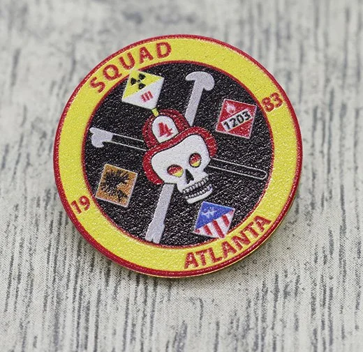 A round service pin with a yellow border and black center, featuring a skull with a red military helmet, surrounded by military-themed patches and text that reads 'SQUAD ATLANTA 1983'.