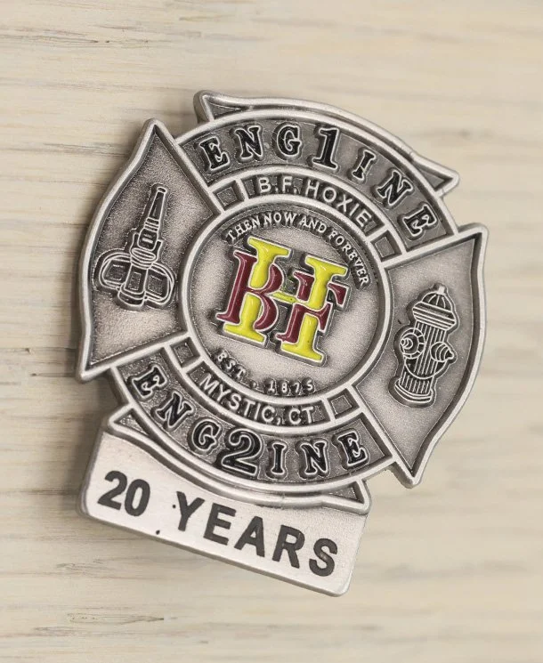 A silver firefighter years of service pin commemorating 20 years, featuring the words 'Engine 1', 'Engine 2', 'B.F. Hoxie', and symbols of a fire nozzle and hydrant, with a central emblem displaying the initials 'K F' in red and yellow.