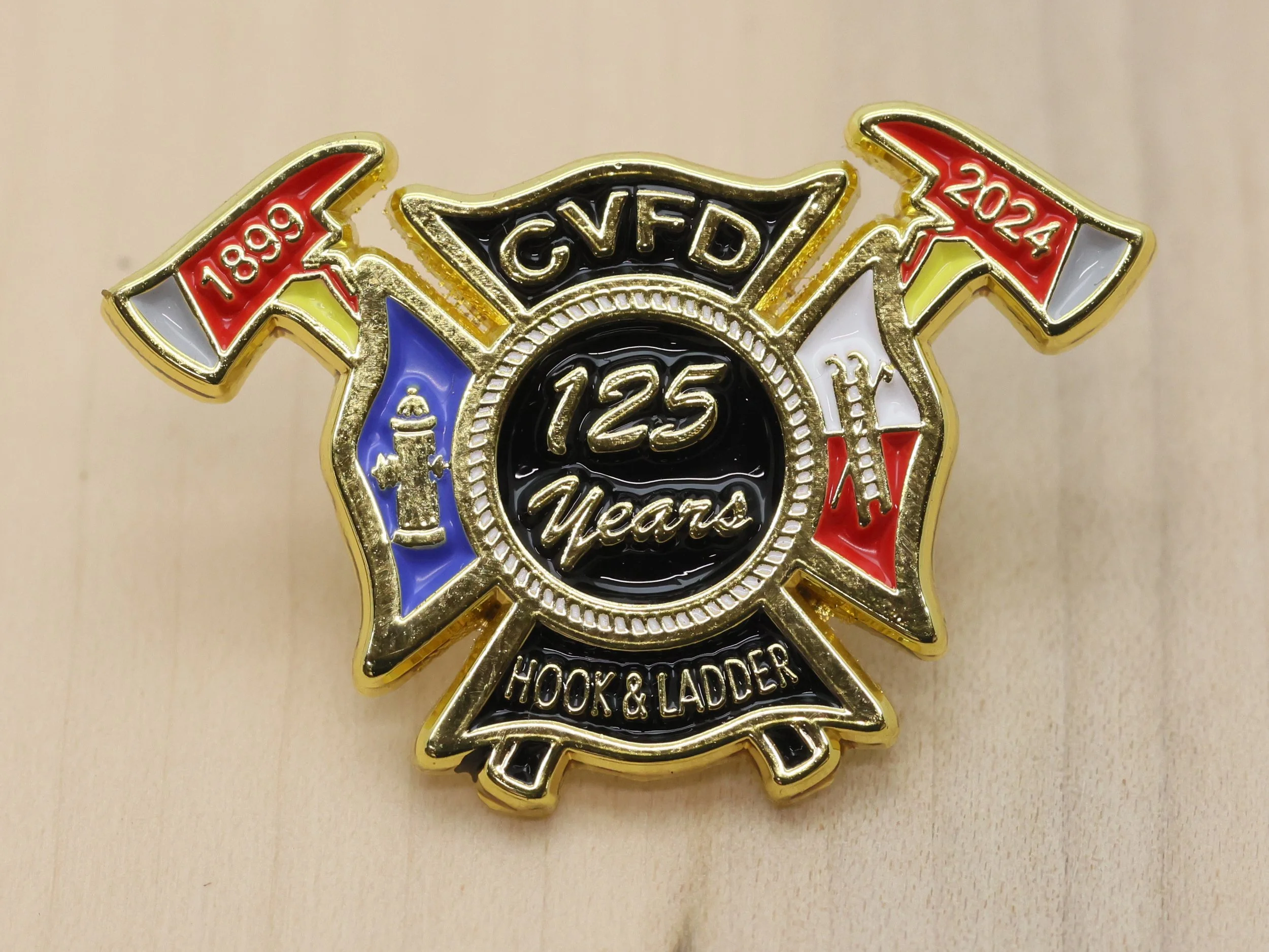 A decorative pin commemorating 125 years of Hook & Ladder. The badge features two crossed red and white flags with years '1899' and '2024' on them, a black central circle with gold text, and additional gold accents.
