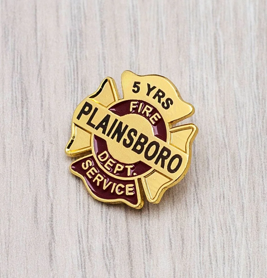 A maltese style pin with the text "5 YRS FIRE PLAINSBORO DEPT. SERVICE" on a wooden surface.