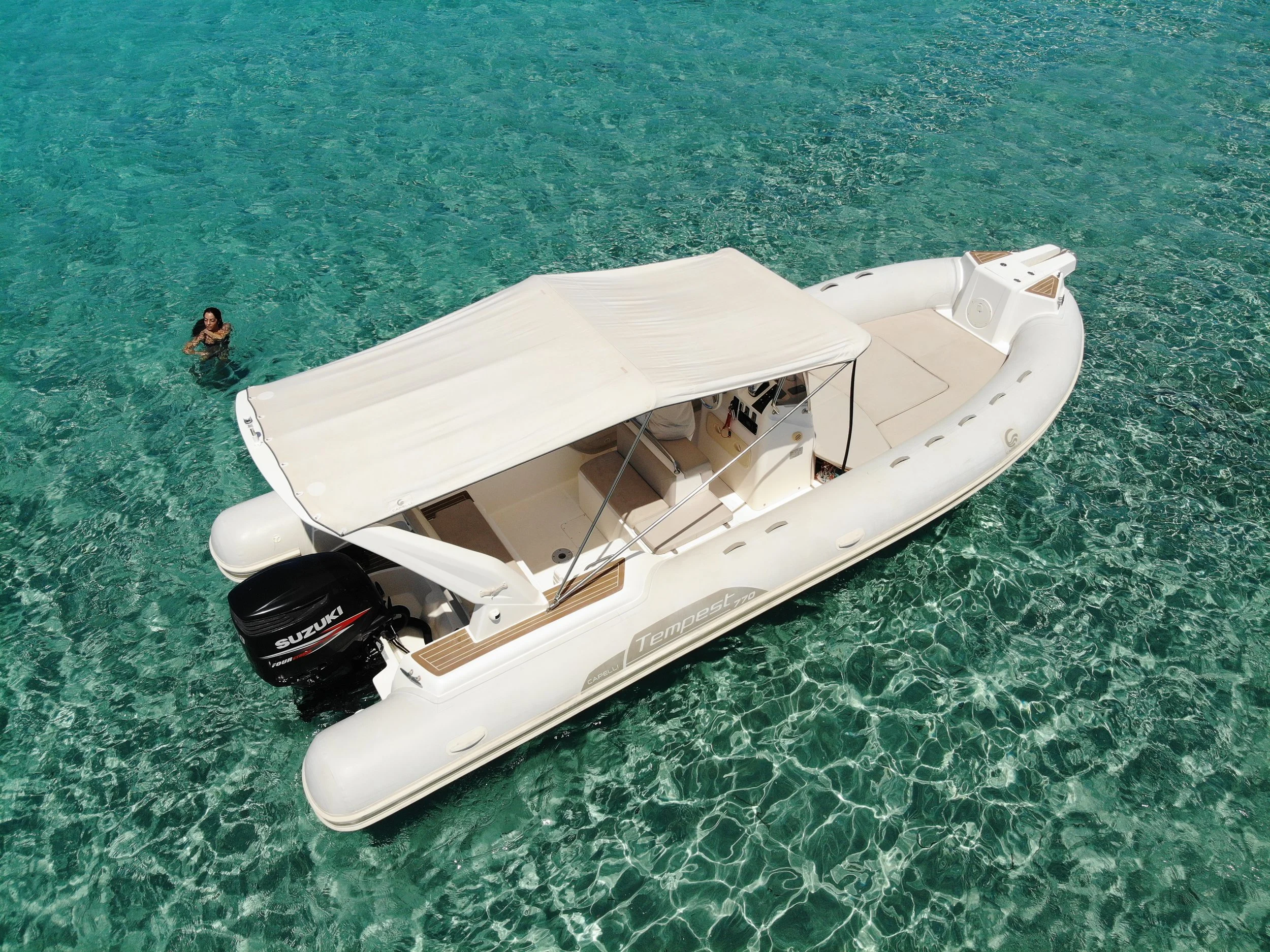 A small white boat with a canopy and an outboard motor floating on clear turquoise water, with a woman swimming nearby.