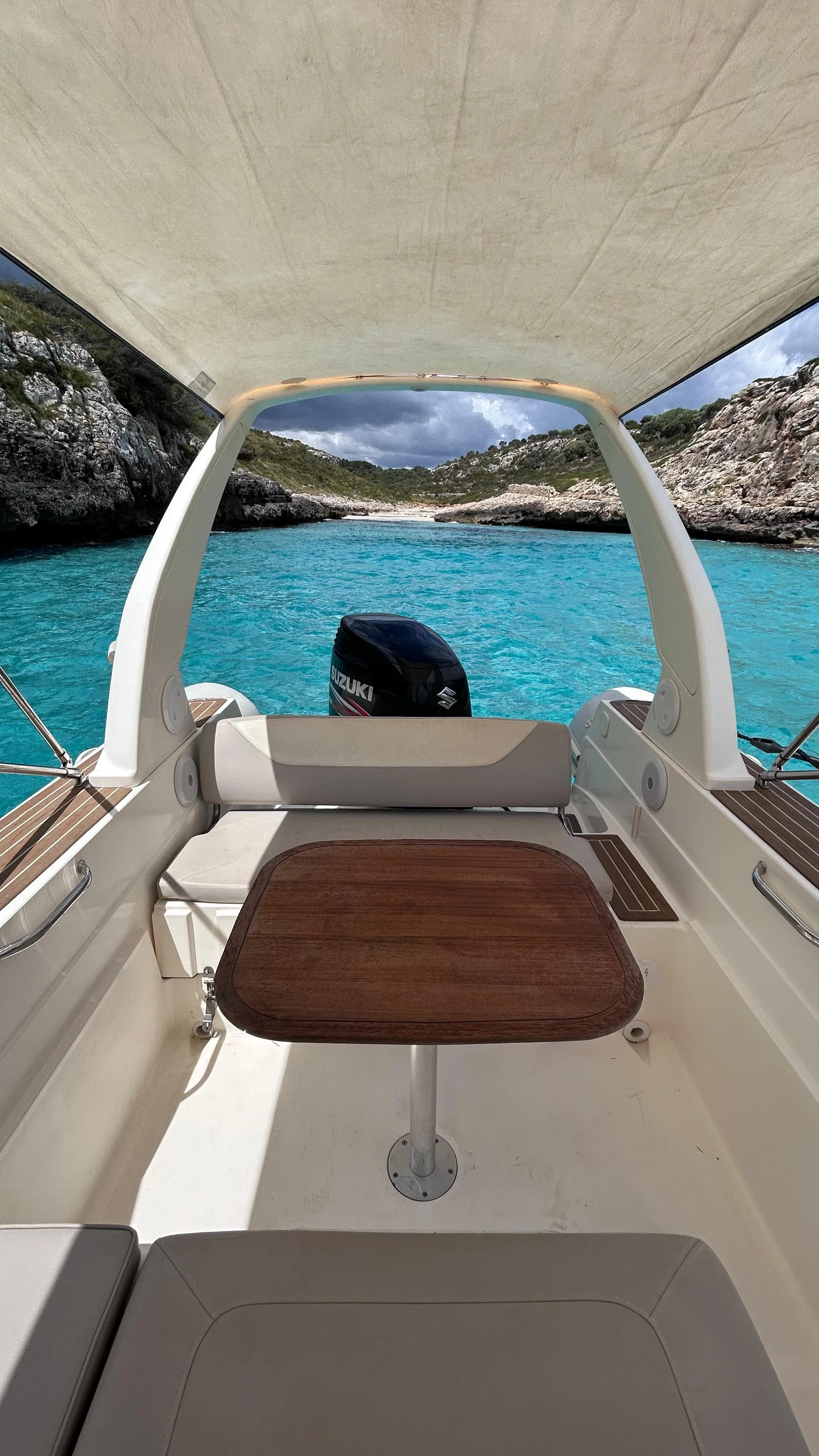 View from the cockpit of a boat looking out over turquoise water with rocky cliffs and cloudy sky in the distance.