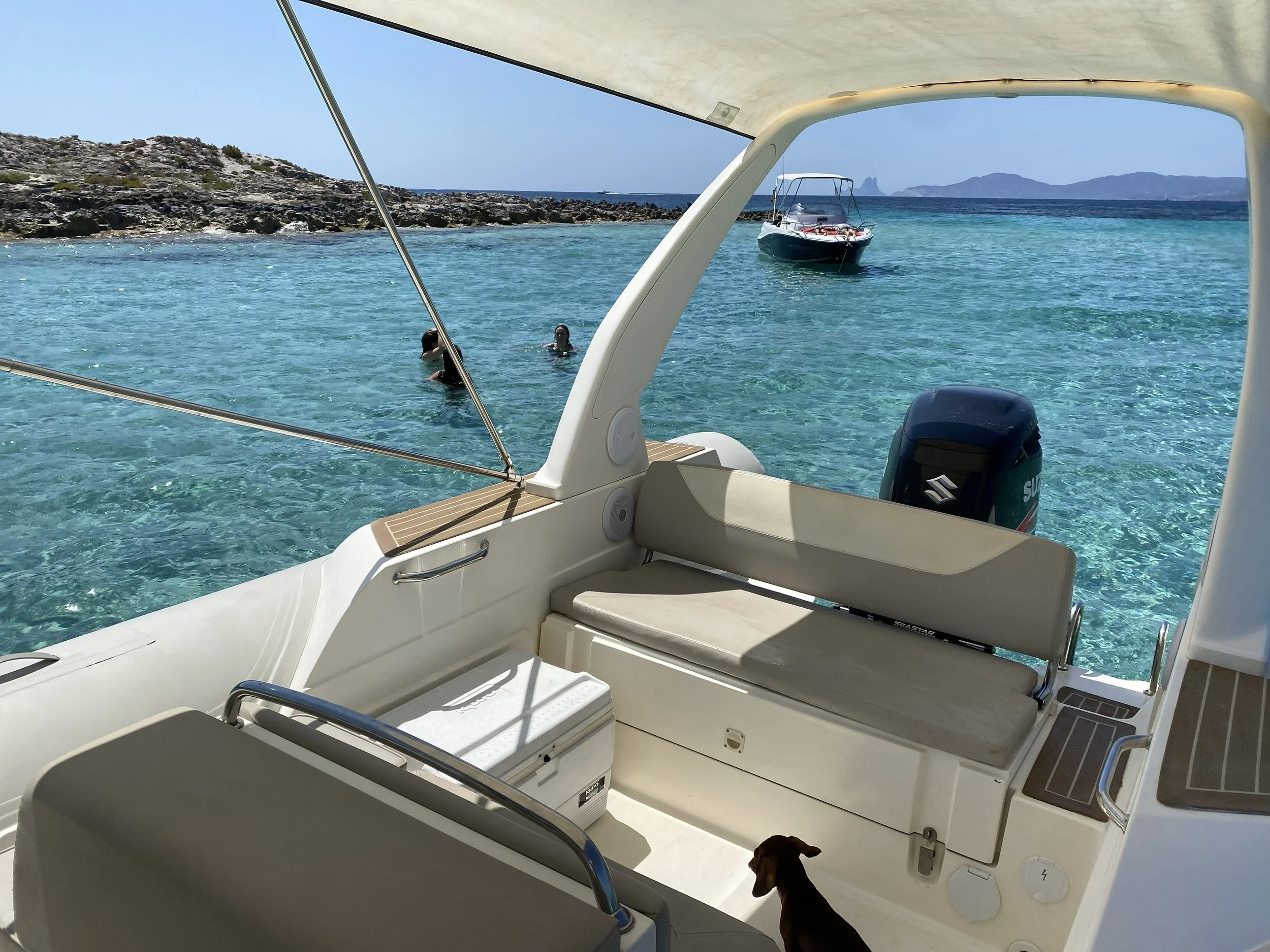 View from a boat showing two people swimming in clear blue water near a rocky shoreline, with a small motorboat anchored nearby and distant islands on the horizon.
