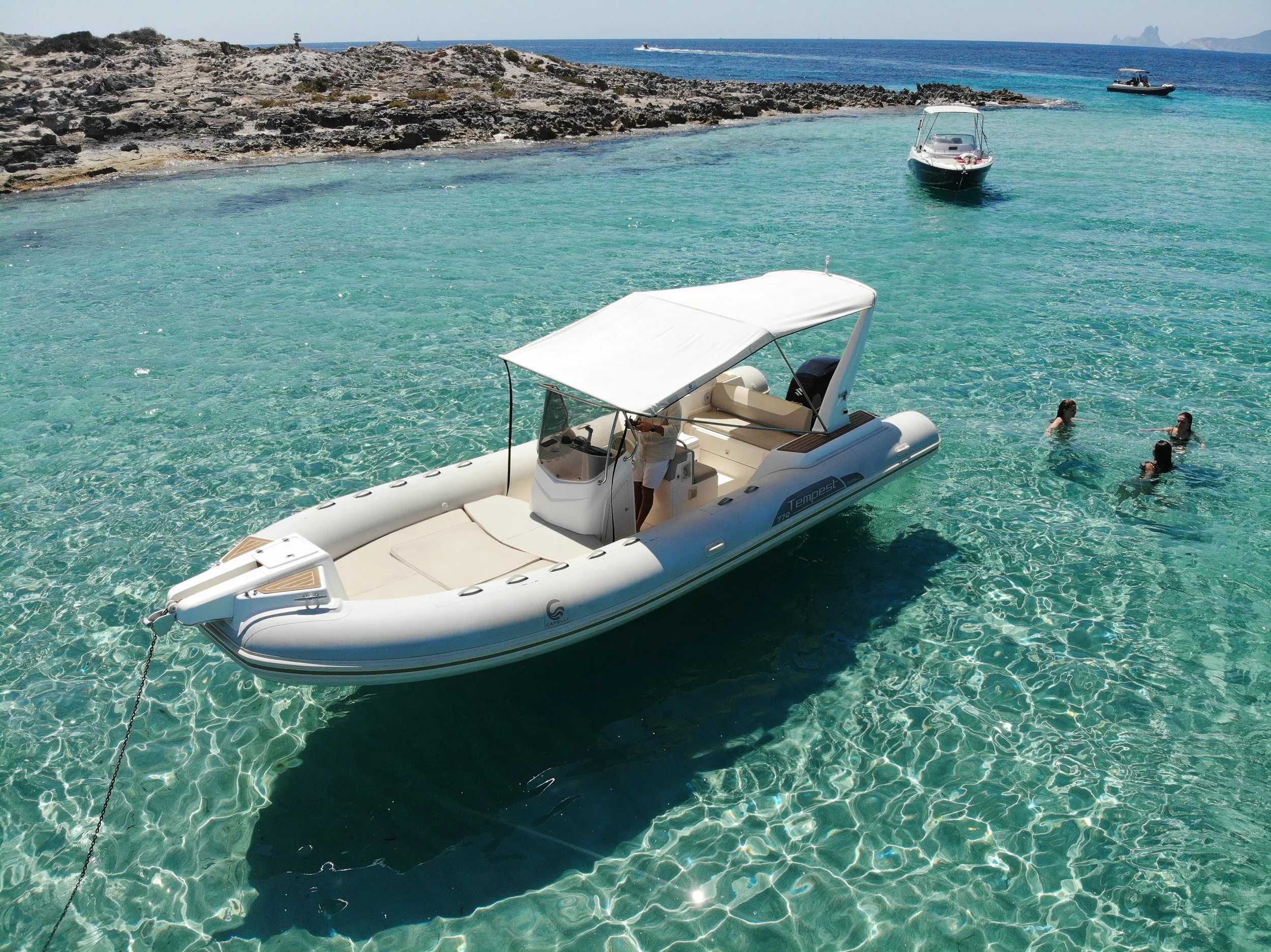 A docked inflatable boat with a canopy floating in clear turquoise water near a rocky shoreline. Three women are swimming in the water nearby, and there are two boats in the distance on the open sea.