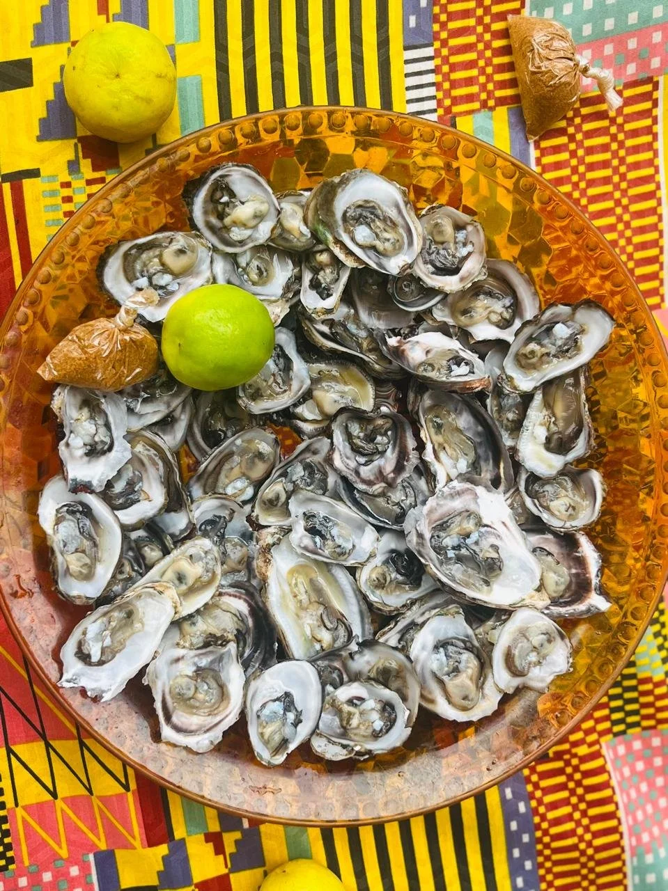 A glass bowl filled with raw oysters on the half shell, a green lime, and two small wrapped candies, placed on a colorful patterned tablecloth.