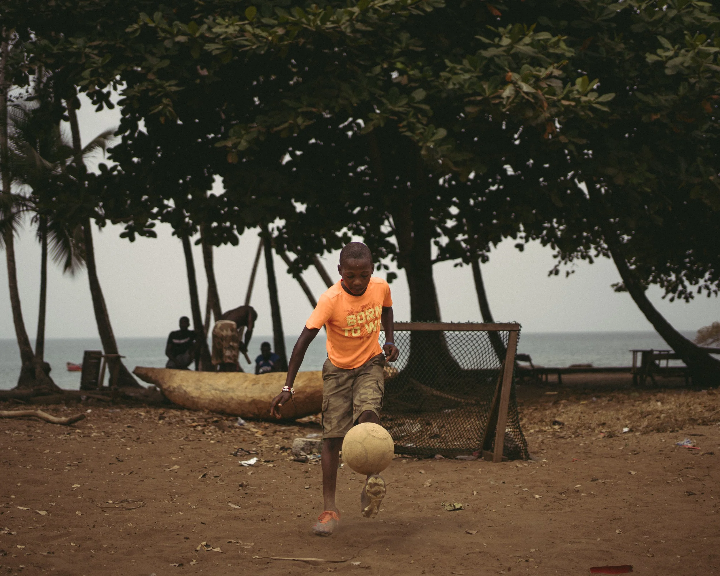 A young boy wearing an orange T-shirt and shorts playing soccer on a dirt field near a beach with trees and a fence in the background.