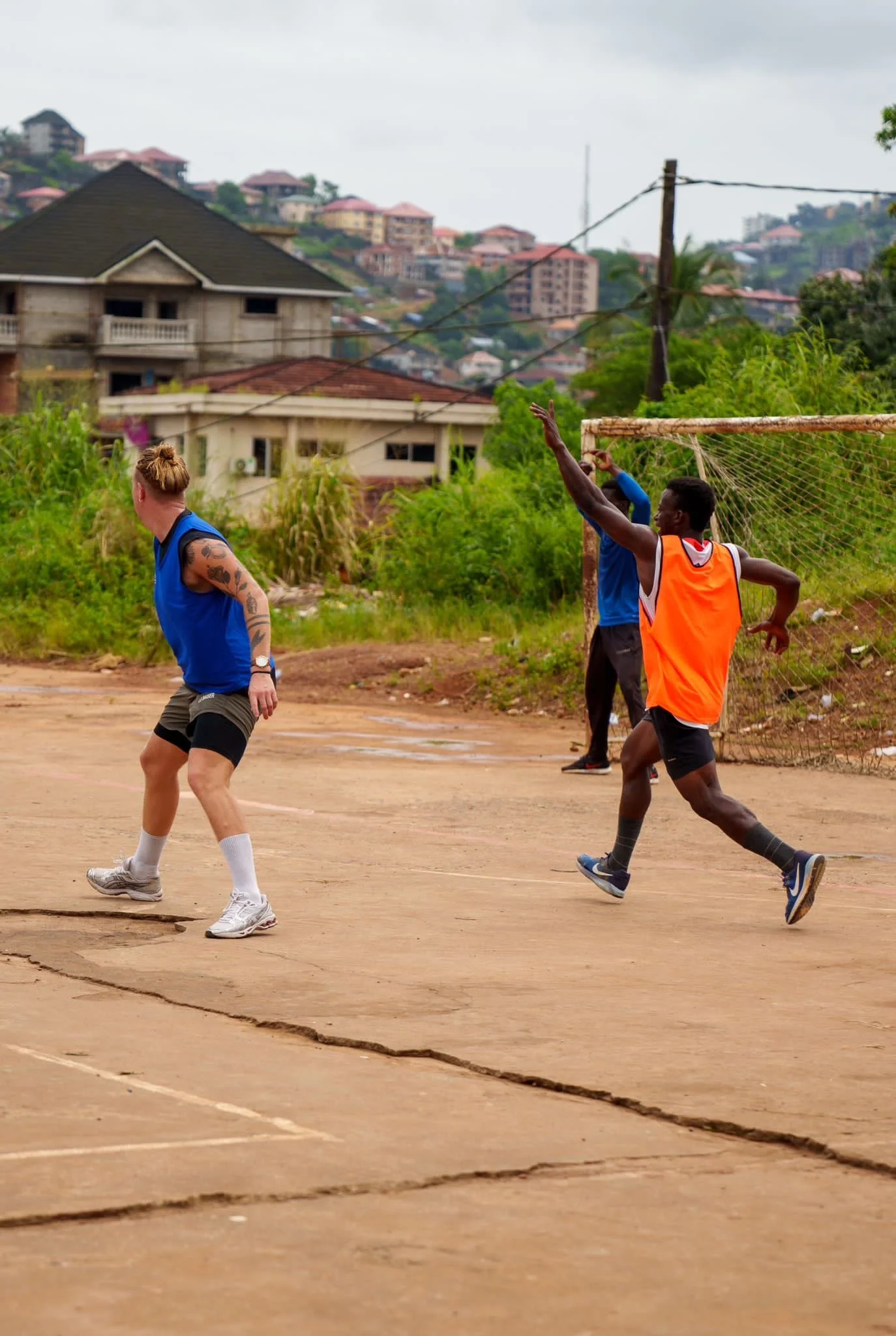 Two runners playing soccer on a dirt field with cracked ground, old goalpost, and a hilly neighborhood with houses in the background.