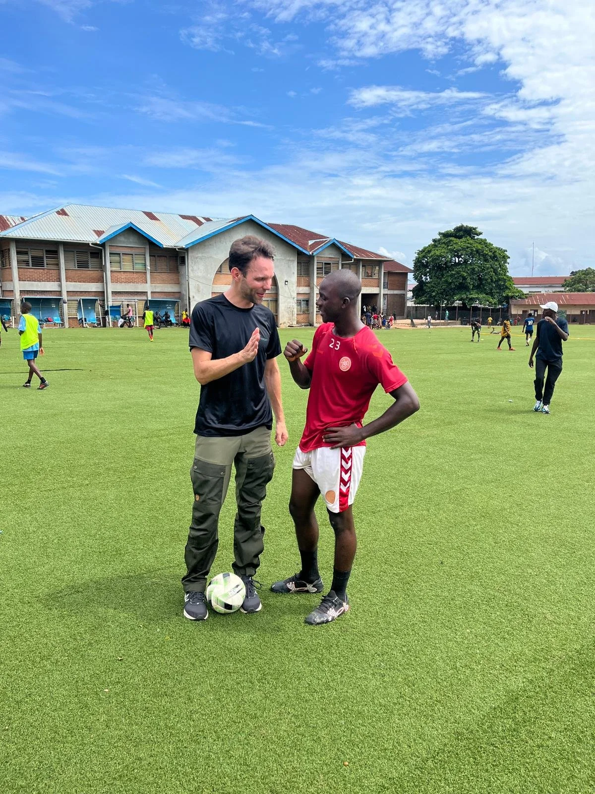 Two men on a soccer field having a conversation, one in a black shirt and green pants, the other in a red sports jersey with the number 23, with a soccer ball near his feet. Others are playing in the background under a partly cloudy sky.