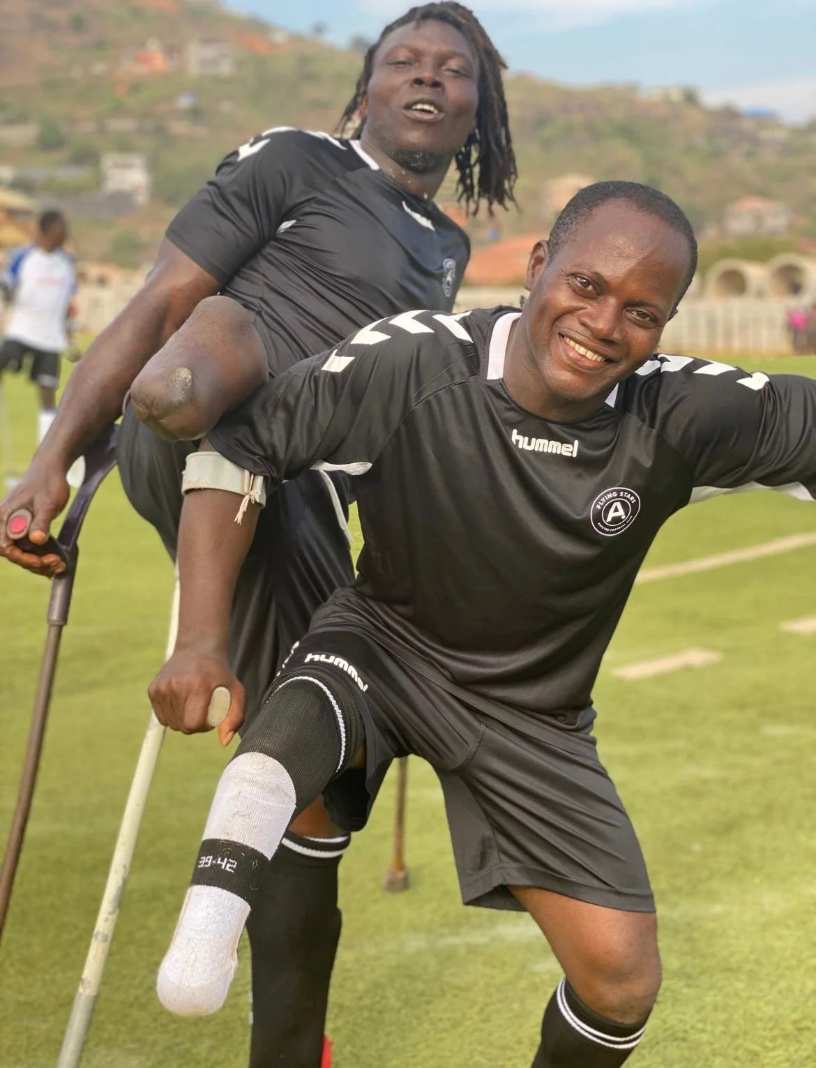Two football players in black jerseys, one with a crutch and a prosthetic leg, celebrating on a grassy field with hills in the background.