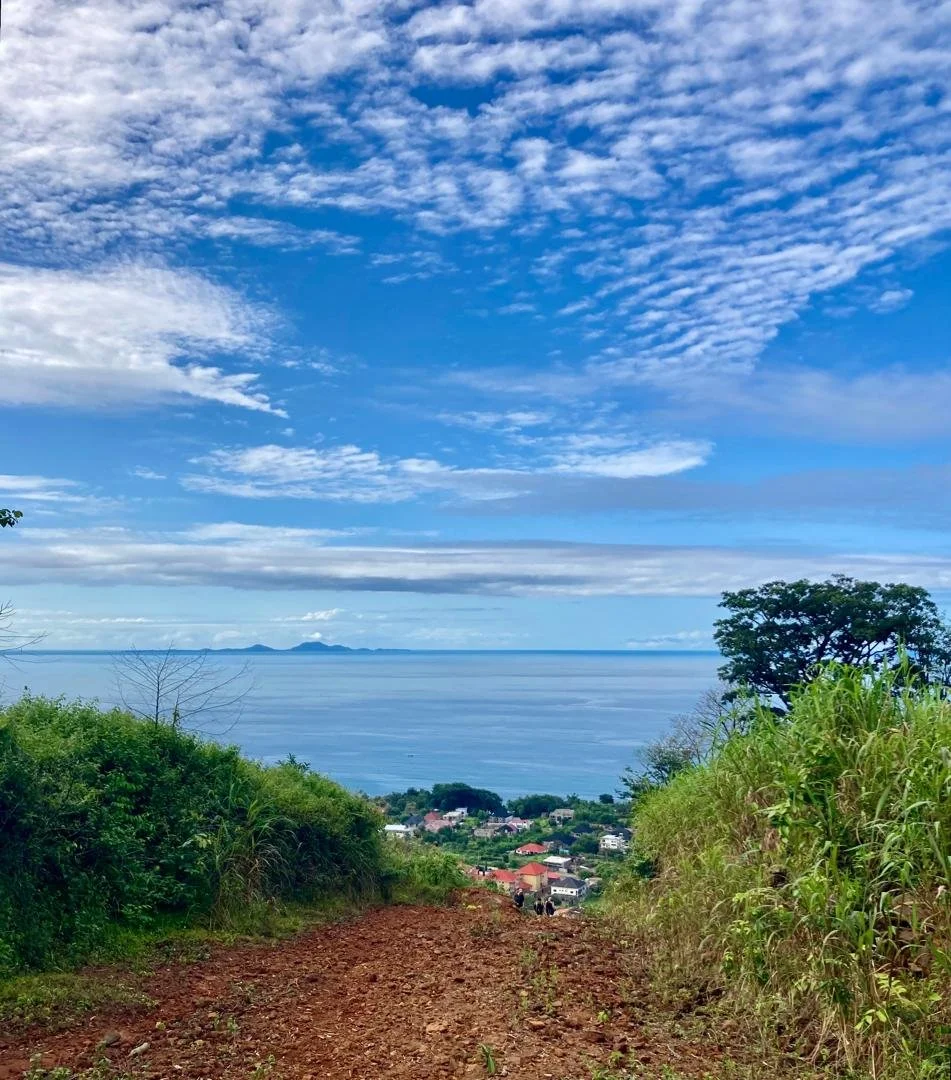A dirt trail leading downhill with greenery on both sides, overlooking a coastal village with houses, the ocean, and distant islands under a partly cloudy blue sky.