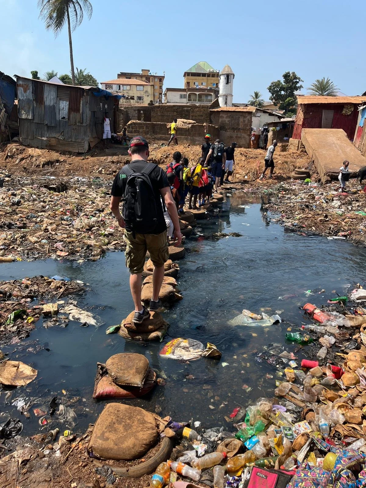 People walking across rocks on a polluted creek filled with trash, including plastic bottles and bags, in an impoverished neighborhood with makeshift homes and taller buildings in the background.