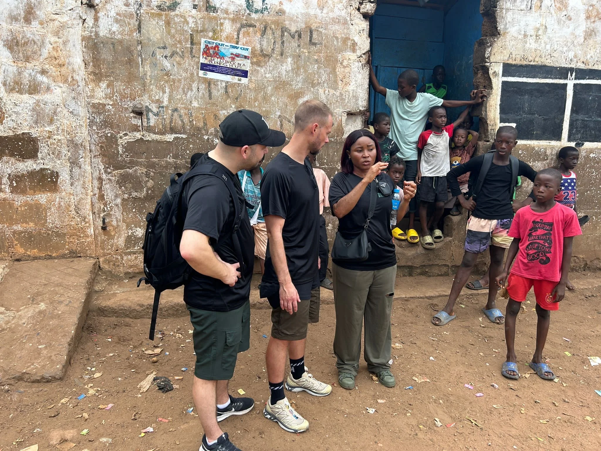 Group of international volunteers and local children standing outside near a weathered brick wall, some children sitting on a step, in a developing community setting.
