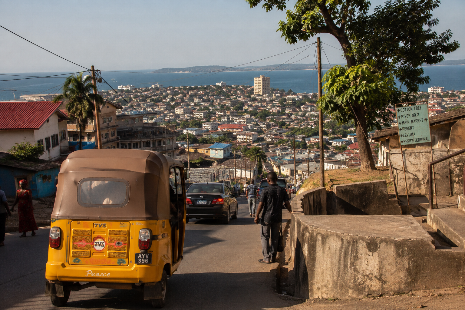 A hilly street in a city with cars and people, overlooking a body of water and buildings in the background.