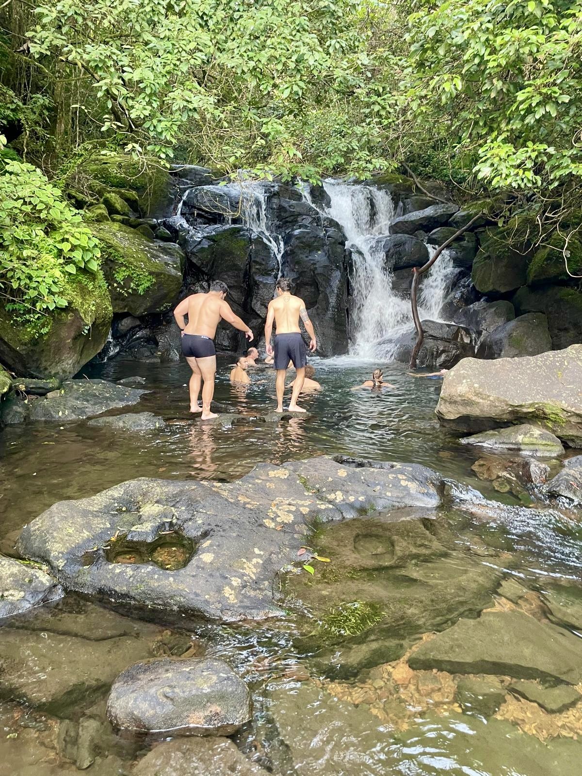 People swimming and wading near a small waterfall in a lush, green forest with rocks and flowing water.