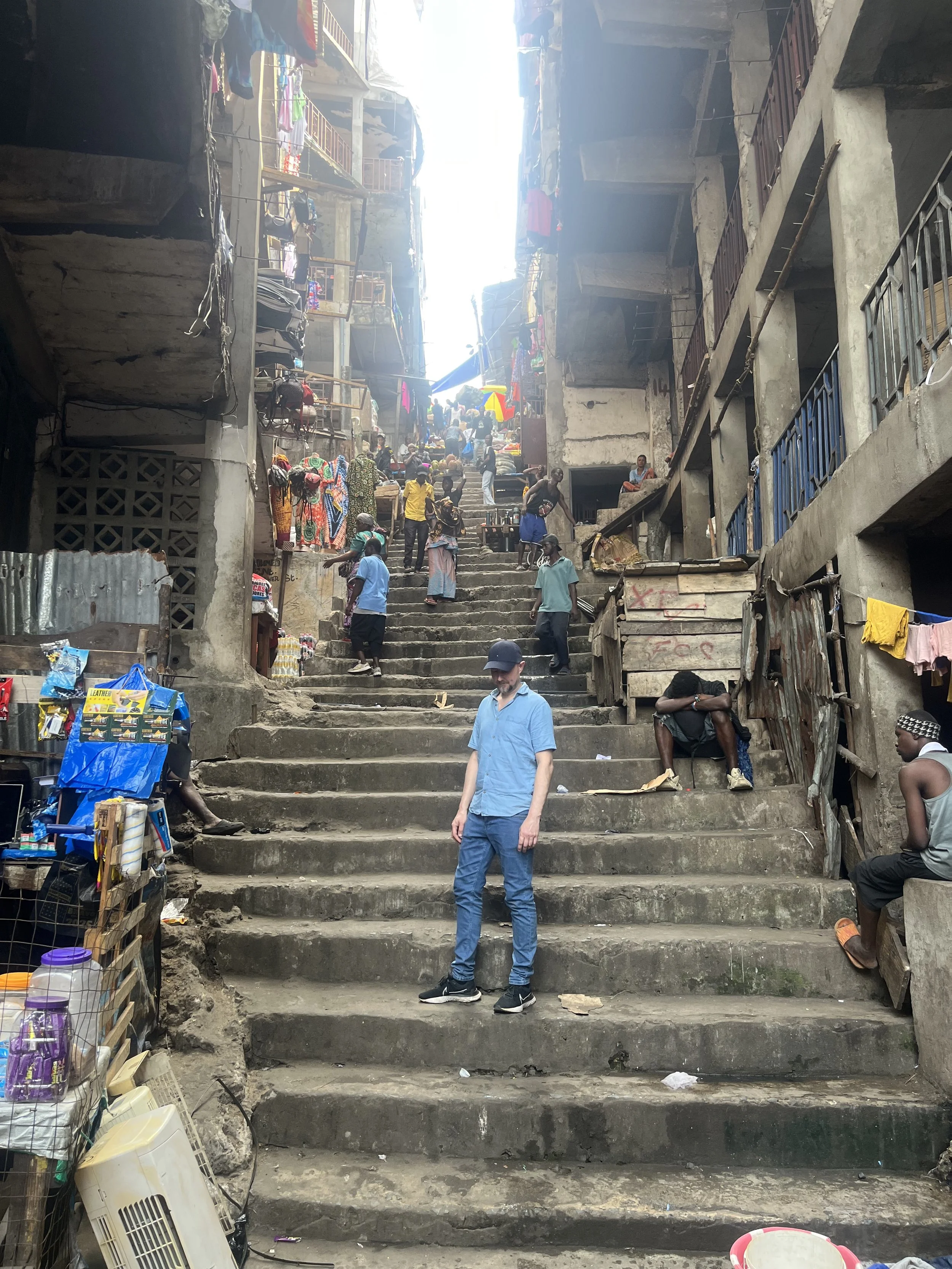 Urban marketplace with people walking and shopping on concrete stairs amidst buildings with balconies, hanging laundry, and street vendors, under a partly cloudy sky.