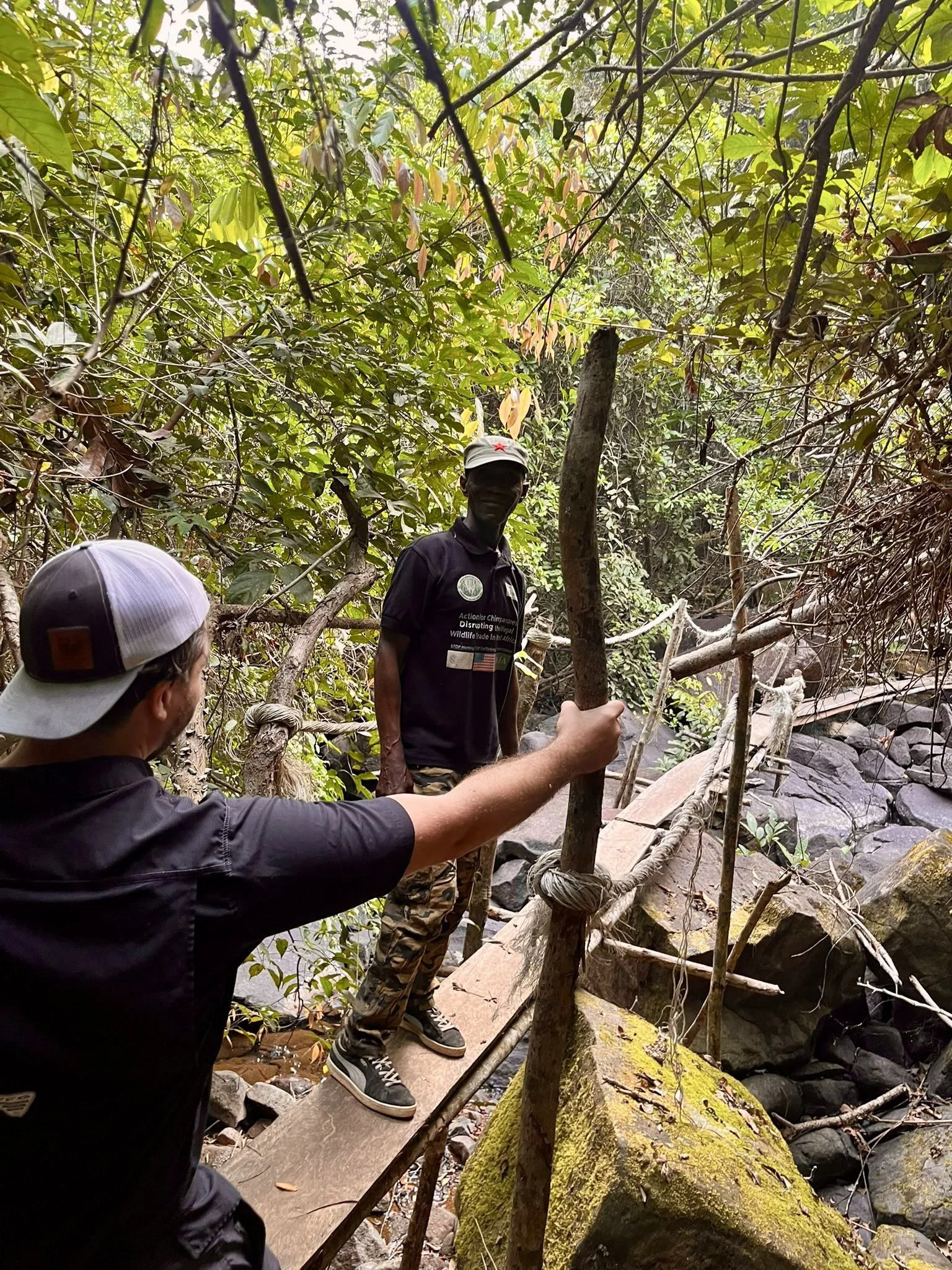 Group of people exploring a lush, wooded area with a small stream, crossing a makeshift wooden bridge over rocks and fallen branches.